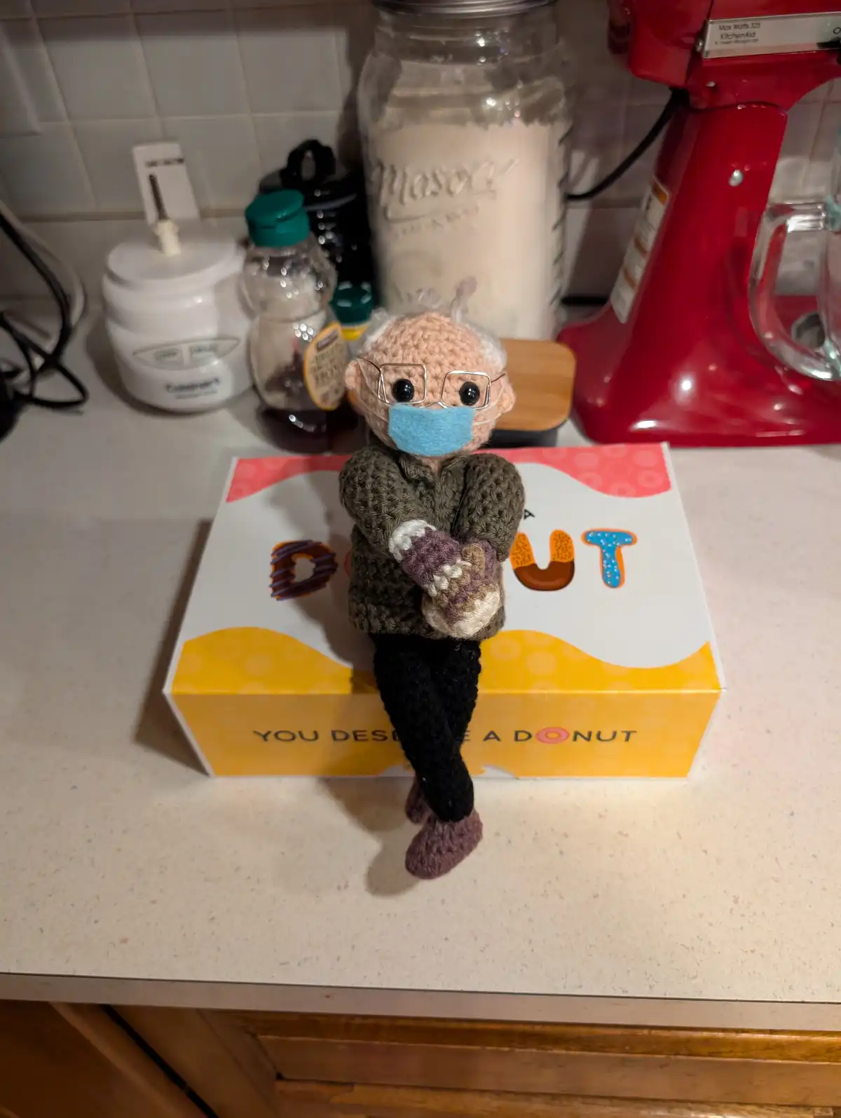 A crocheted elderly doll with glasses and a blue face mask sits on a donut box that reads 'YOU DESERVE A DONUT'. The doll…
