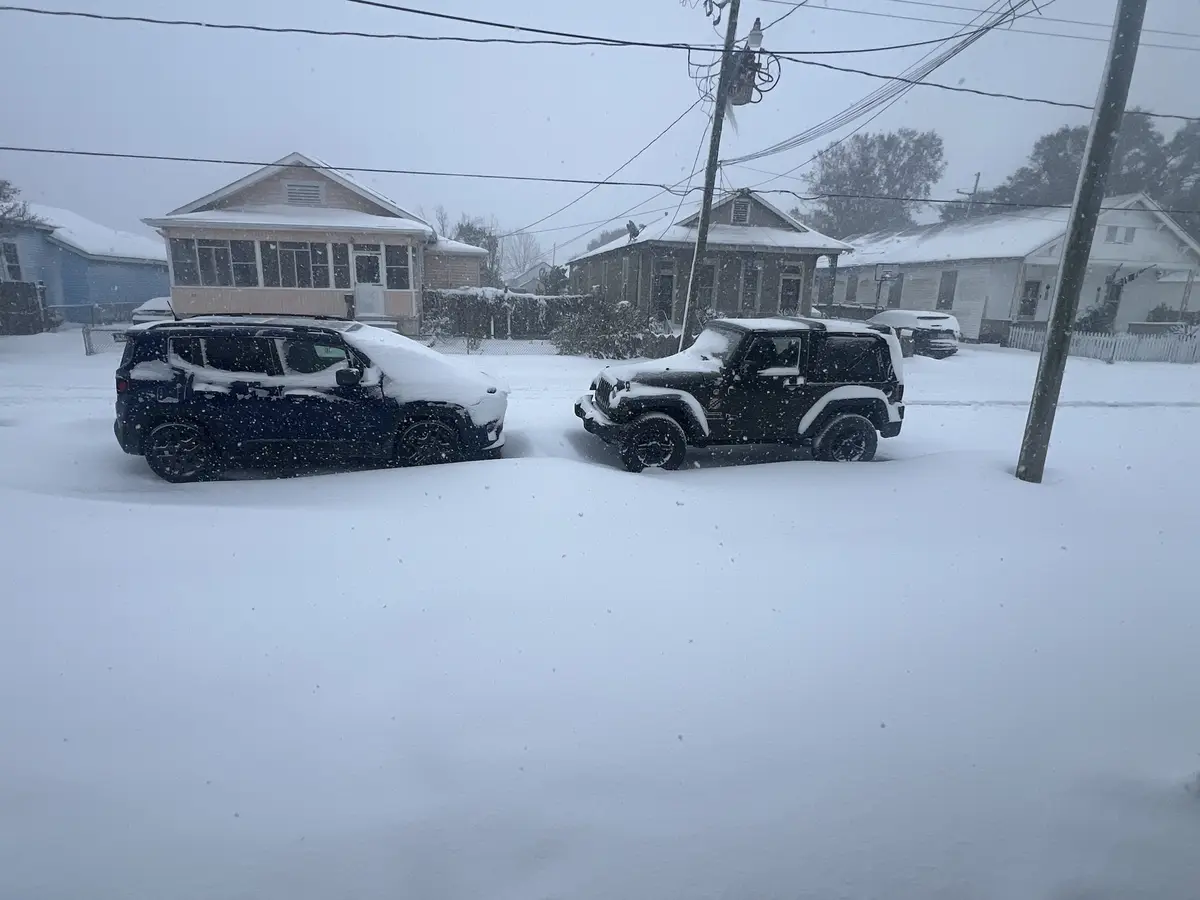 Two cars, a dark SUV and a black Jeep Wrangler, are parked side-by-side in a residential street completely buried under deep…