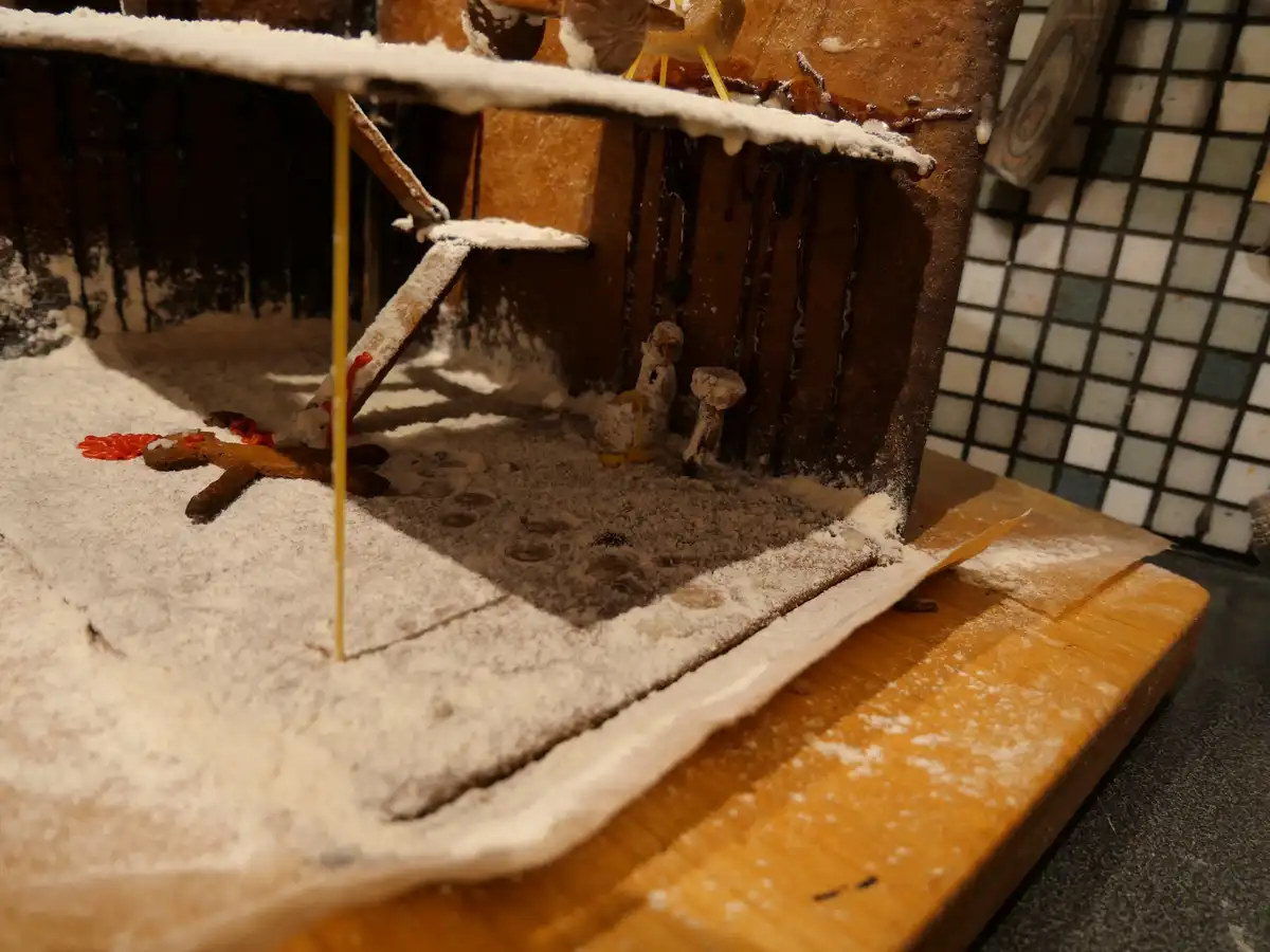 A close-up of a poorly constructed gingerbread house covered in powdered sugar, with melted icing dripping down the sides…