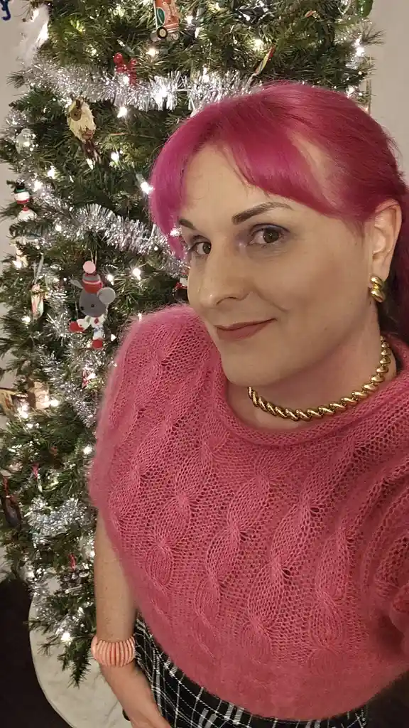 A woman with bright pink hair and a matching pink sweater stands in front of a decorated Christmas tree with lights and…