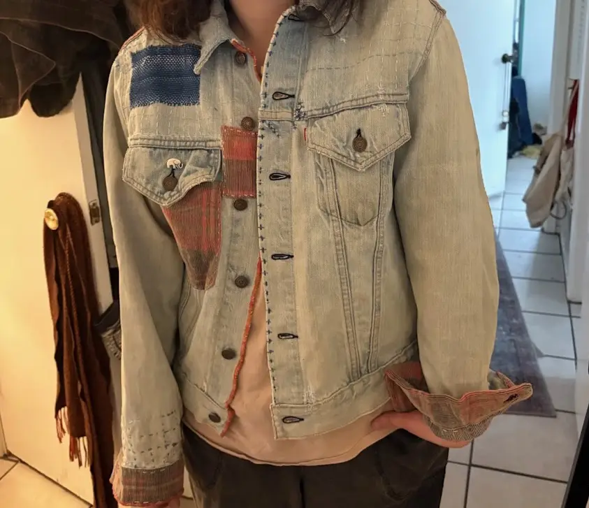 A person is wearing a heavily patched and customized denim jacket with visible fabric repairs, including a blue woven patch…