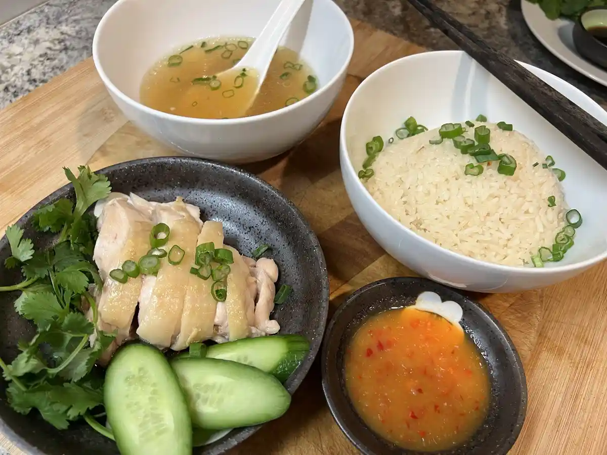 A photo of a home-cooked meal featuring sliced chicken with cilantro and cucumber in a dark bowl, a bowl of white rice…