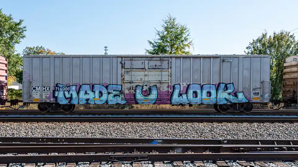 A silver freight train car sits on railway tracks under a clear blue sky, with trees in the background. The side of the car…