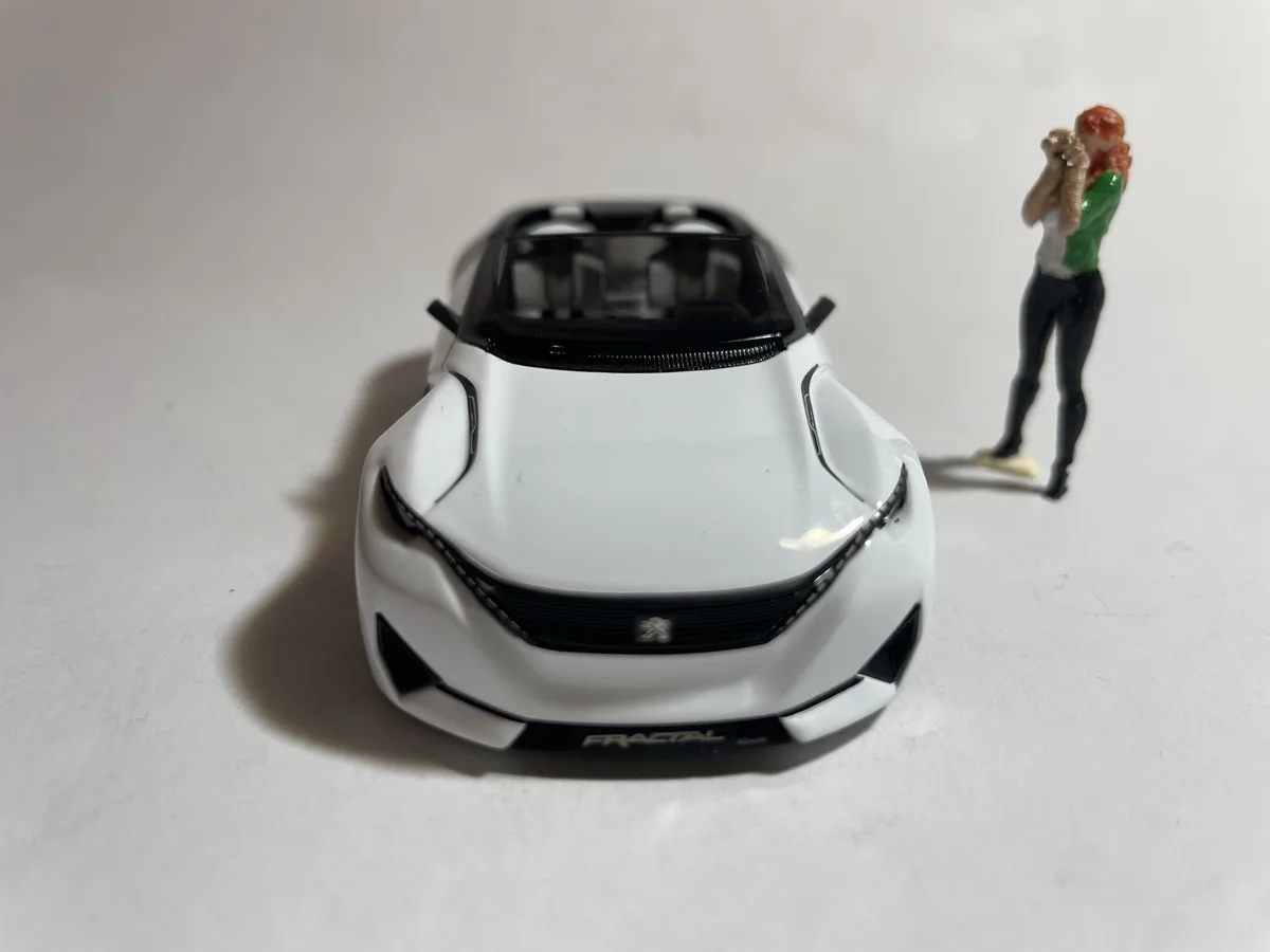 A white toy car, identified as a Peugeot Fractal model, is positioned on a plain white surface. To its right stands a small…