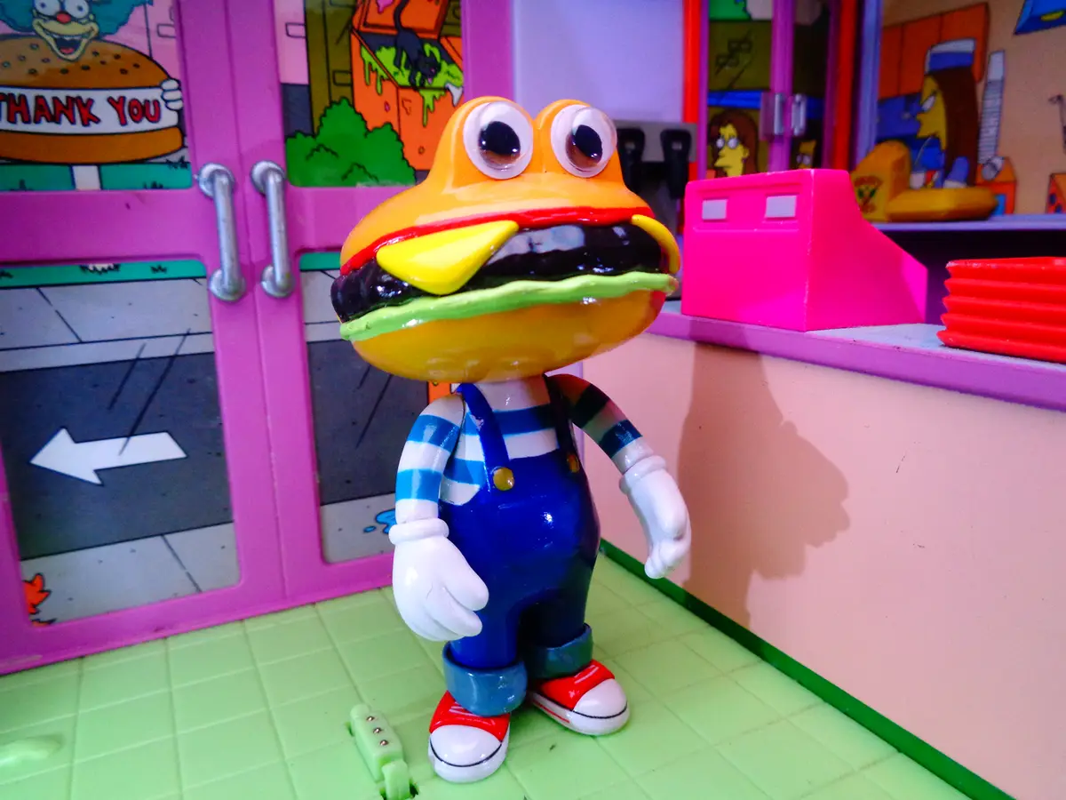A toy figure with a hamburger-shaped head stands in a colorful, cartoonish playset resembling a fast-food restaurant. The…