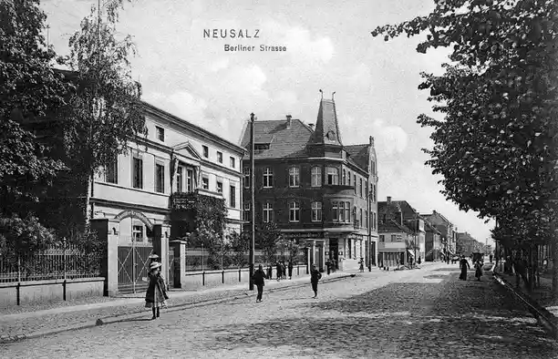 A vintage black-and-white postcard of Neusalz, Berliner Strasse, showing cobblestone streets, period architecture with…