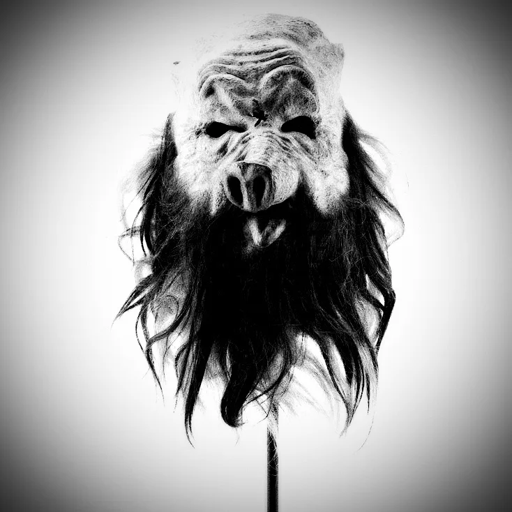 A black-and-white image shows a grotesque, wrinkled mask with a pig-like snout and long, shaggy hair, mounted on a stand…