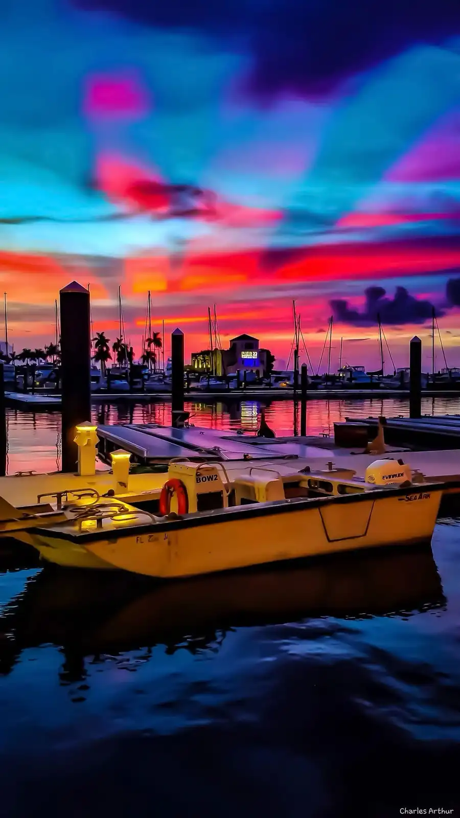 A vibrant, heavily saturated sunset over a marina with boats docked, featuring a yellow skiff in the foreground. The sky is…