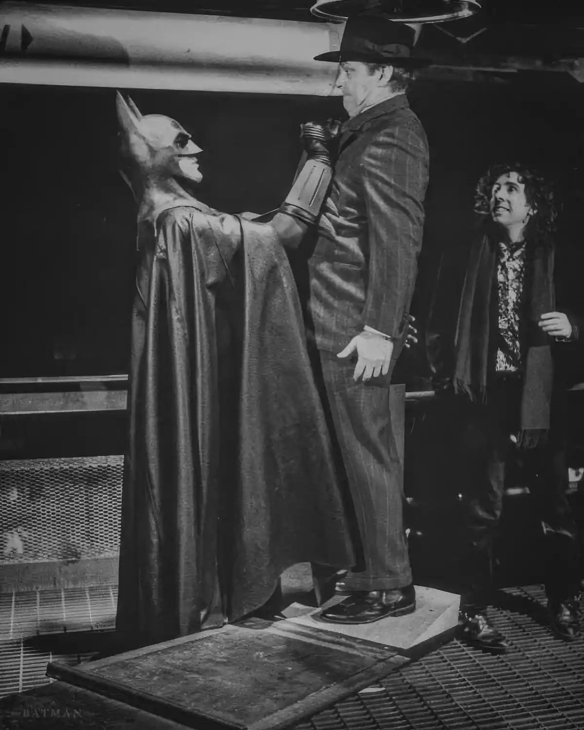 A black-and-white photo shows a person in a vintage Batman costume with a cape and cowl, standing on a platform and holding…