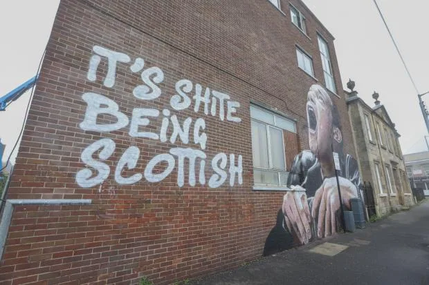 A large mural on a brick building shows a person screaming in anguish while the text 'IT'S SHITE BEING SCOTTISH' is painted…
