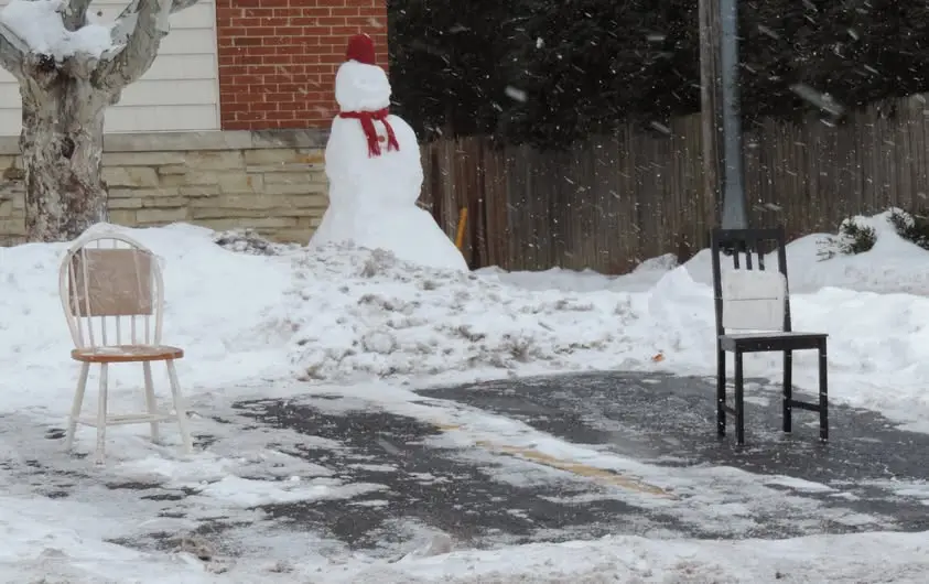 A snowy yard shows a snowman wearing a red hat and scarf standing beside two chairs—one white and one black—placed on a…