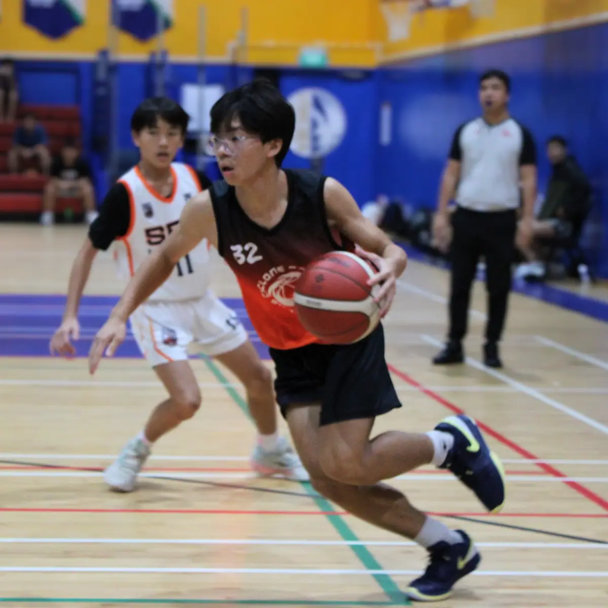 A young male basketball player in a black and orange jersey with the number 32 is dribbling a basketball while running down…
