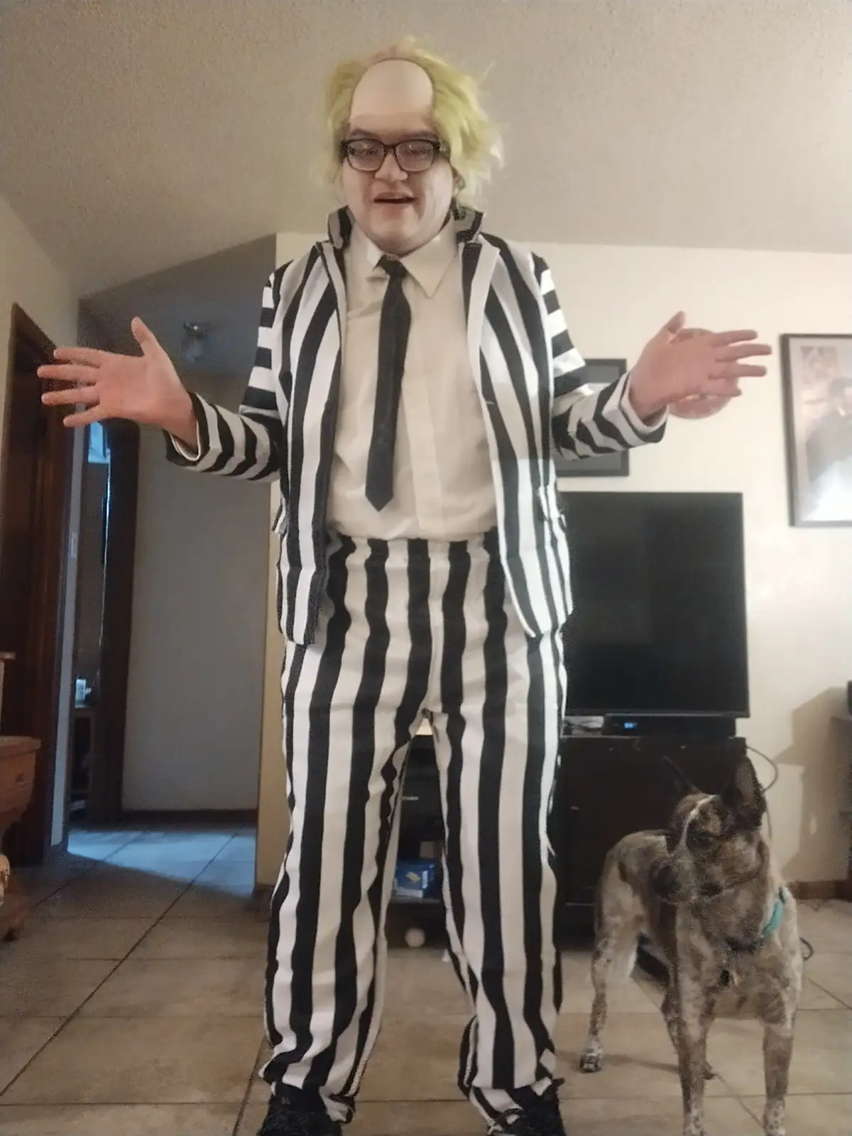 A person in a full Beetlejuice costume, including a bald cap with green wig, glasses, and black-and-white striped suit…