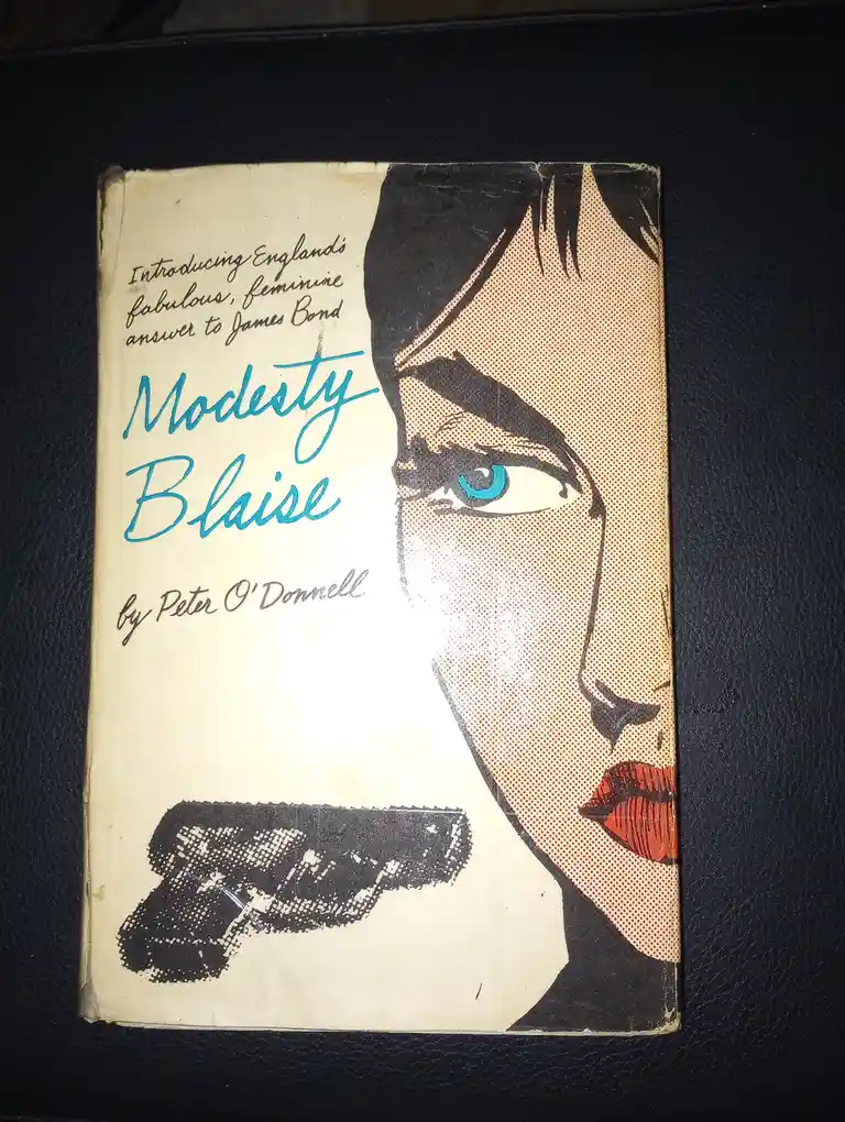 A vintage paperback book cover for 'Modesty Blaise' by Peter O'Donnell, featuring a stylized illustration of a woman's face…