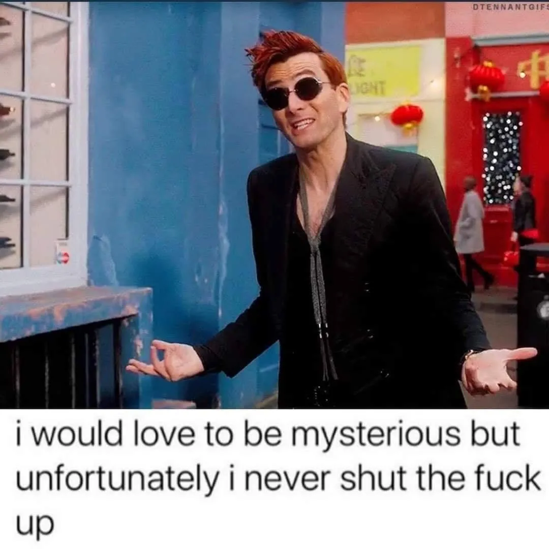 A man with red hair and sunglasses, wearing a black jacket and tie, stands with his hands open in a shrugging gesture. The…