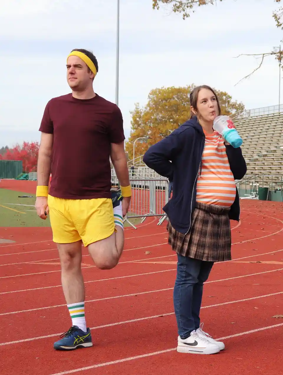 A man in yellow shorts and headband stretches on a red running track while a woman in a plaid skirt and hoodie sips from a…