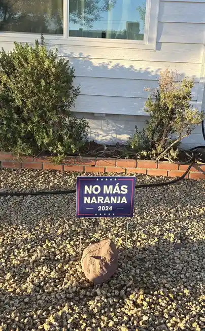 A blue political-style yard sign reading 'NO MÁS NARANJA 2024' stands in gravel in front of a white house, with a large…