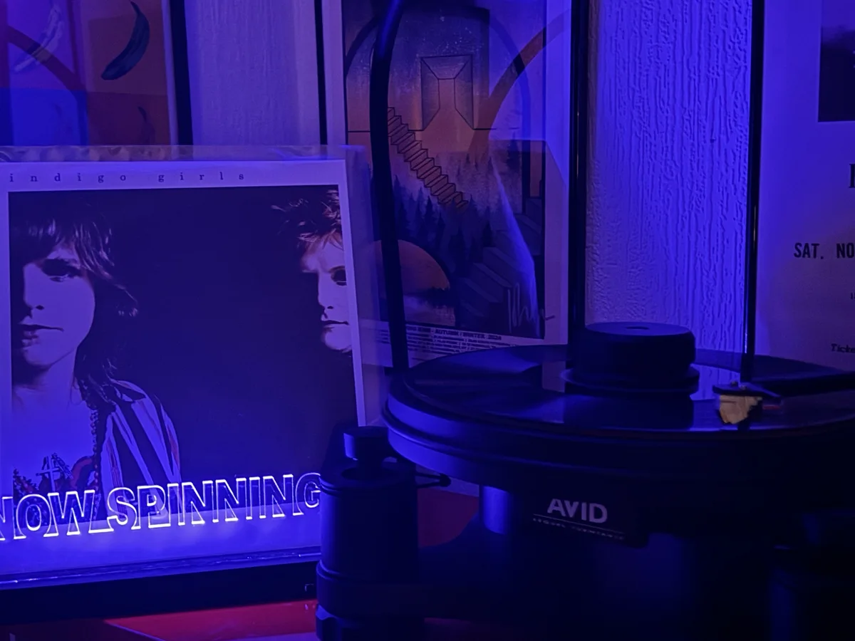 A close-up shot of a black AVID turntable under blue lighting, with a vinyl record on the platter and a tonearm. Behind it…