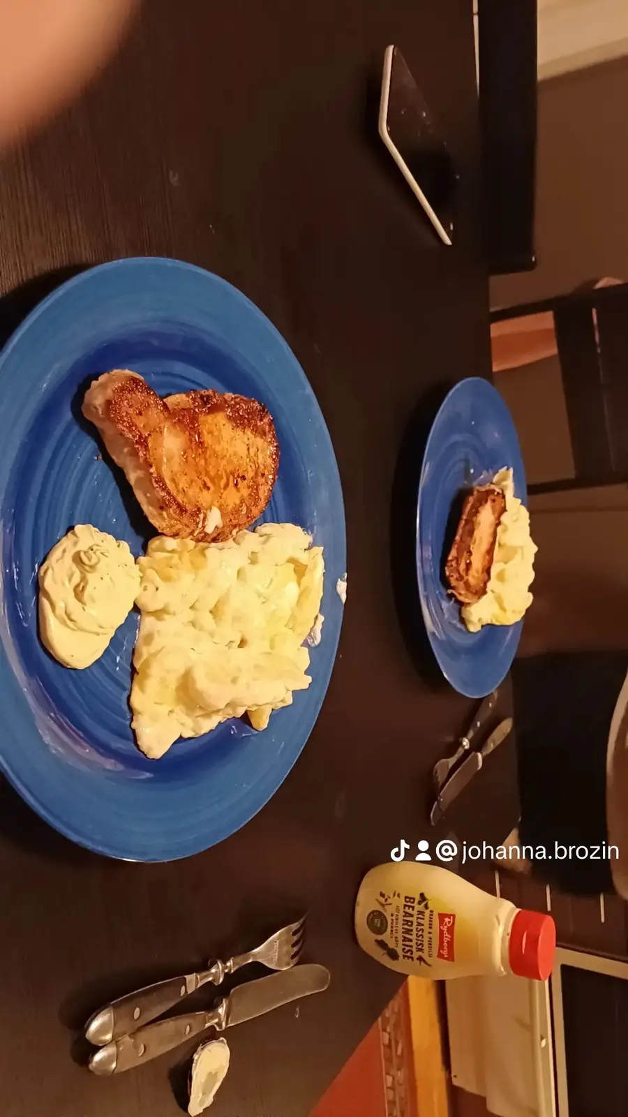 An overhead shot shows two blue plates on a dark table, one with a large piece of seared meat and scrambled eggs, the other…