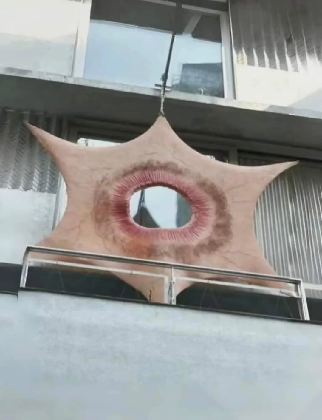 a large, star-shaped, flesh-colored object with a central hole resembling a mouth hangs from a chain above a balcony. The…