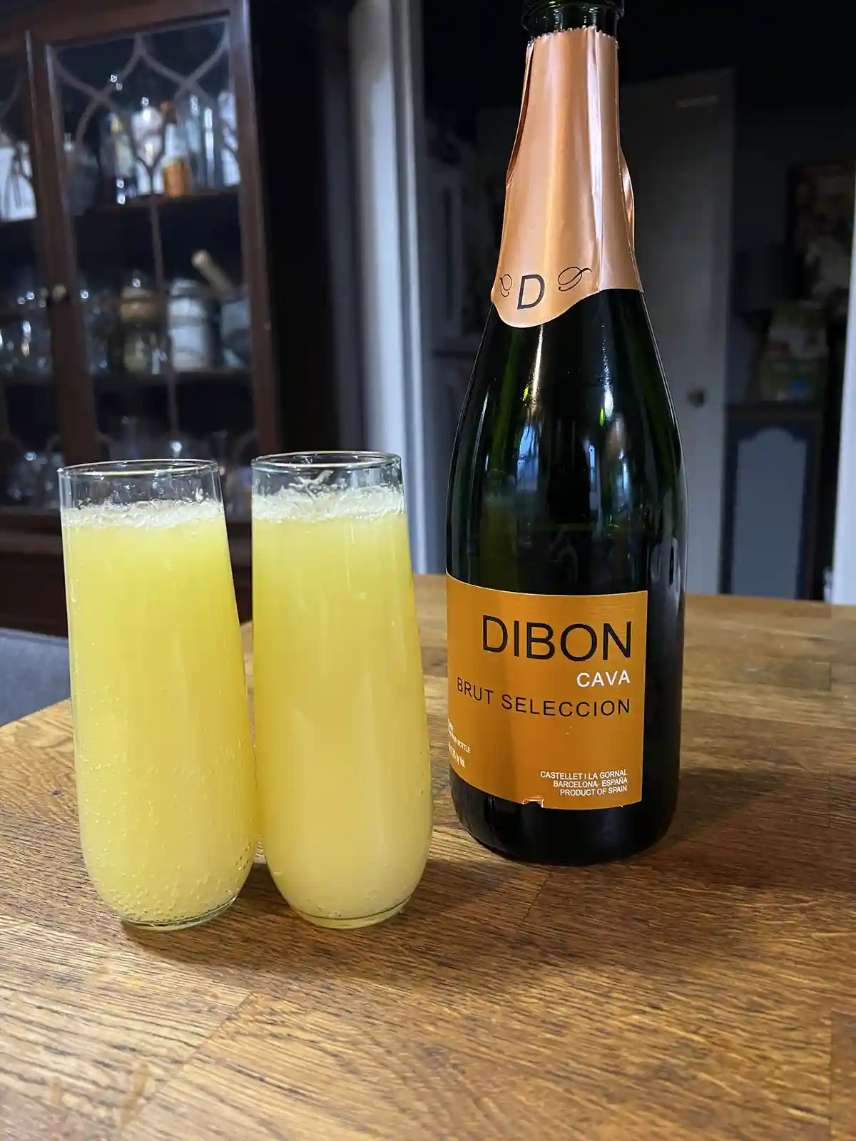 A bottle of Dibon Brut Selección Cava stands next to two tall glasses filled with a bubbly, pale yellow liquid, likely a…