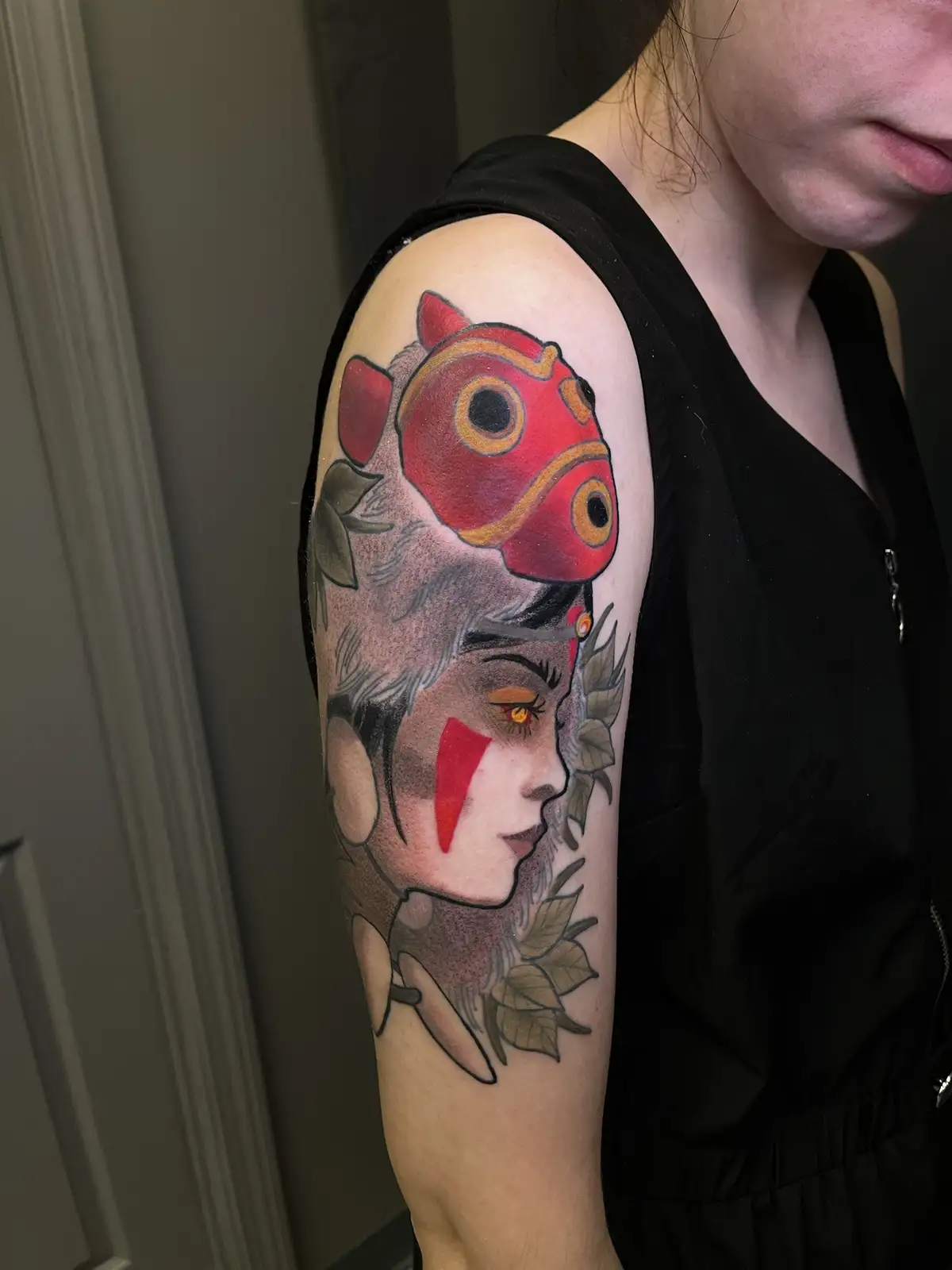A person's upper arm displays a detailed tattoo of a red fish mask with large eyes, positioned above a stylized female face…