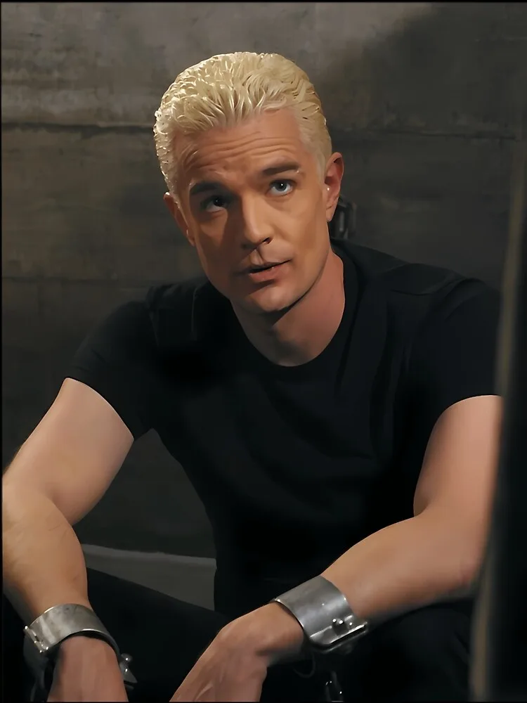 A man with platinum blonde hair and a black t-shirt sits with metal handcuffs on both wrists, looking upward with a confused…