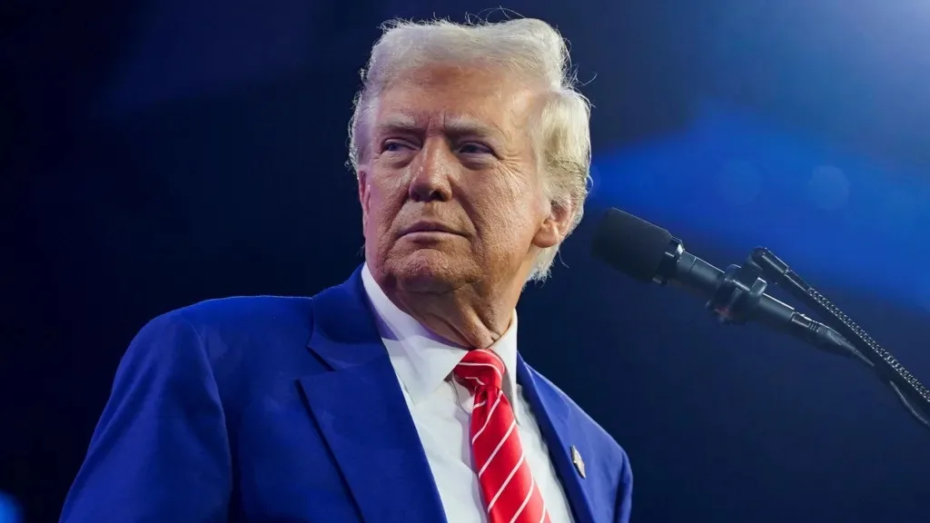 a close-up photo of Donald Trump wearing a blue suit, white shirt, and red tie, looking off to the side with a serious…