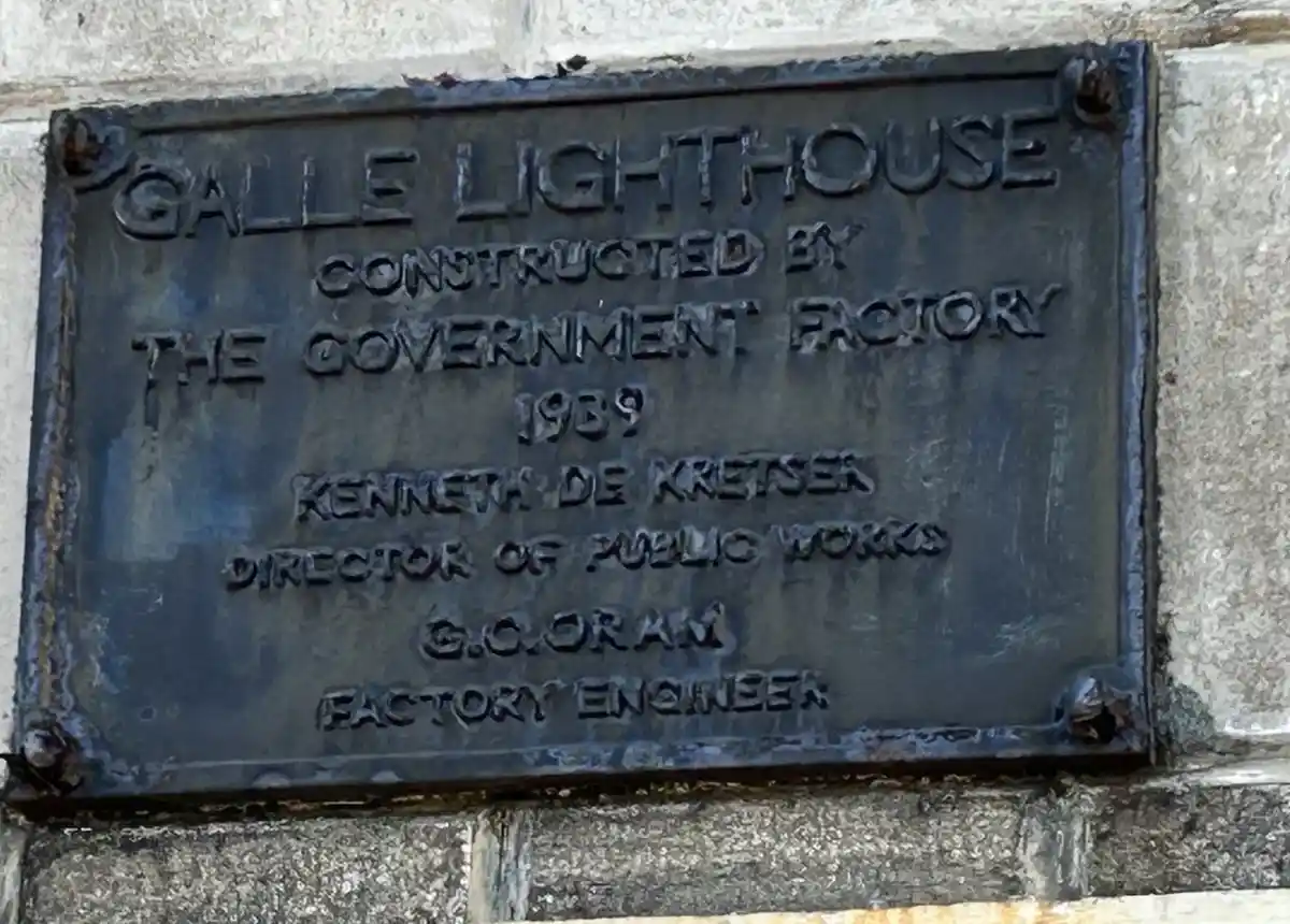 A close-up of a weathered, dark metal plaque mounted on a stone wall, engraved with text identifying the Galle Lighthouse as…