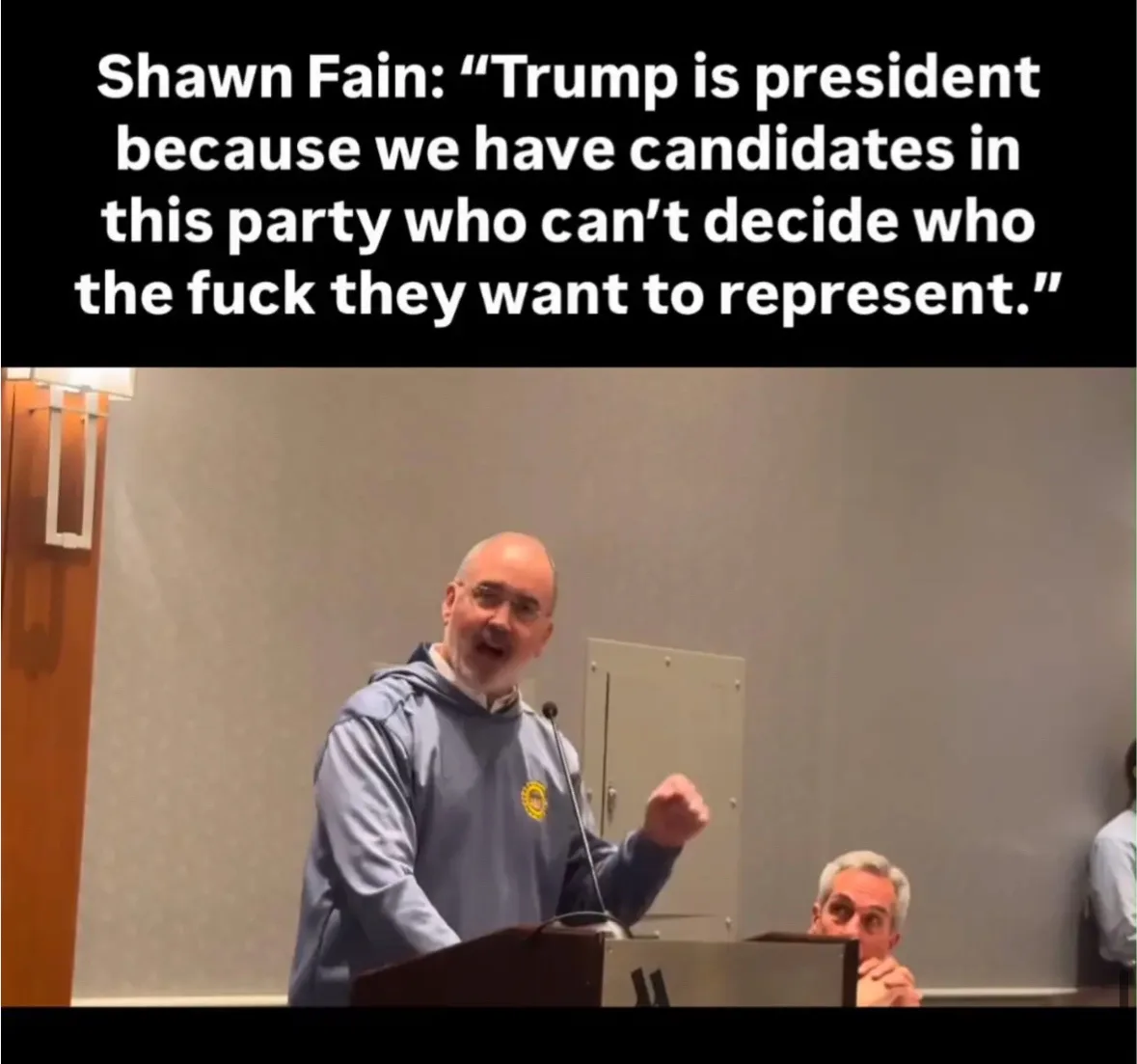 A man in a blue hoodie stands at a podium speaking passionately with his fist raised, while another man sits in the…