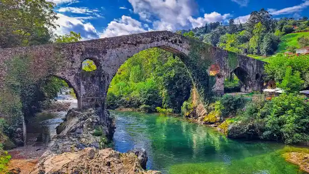A scenic, heavily vine-covered stone arch bridge spans a clear, green river under a bright blue sky with fluffy clouds. The…