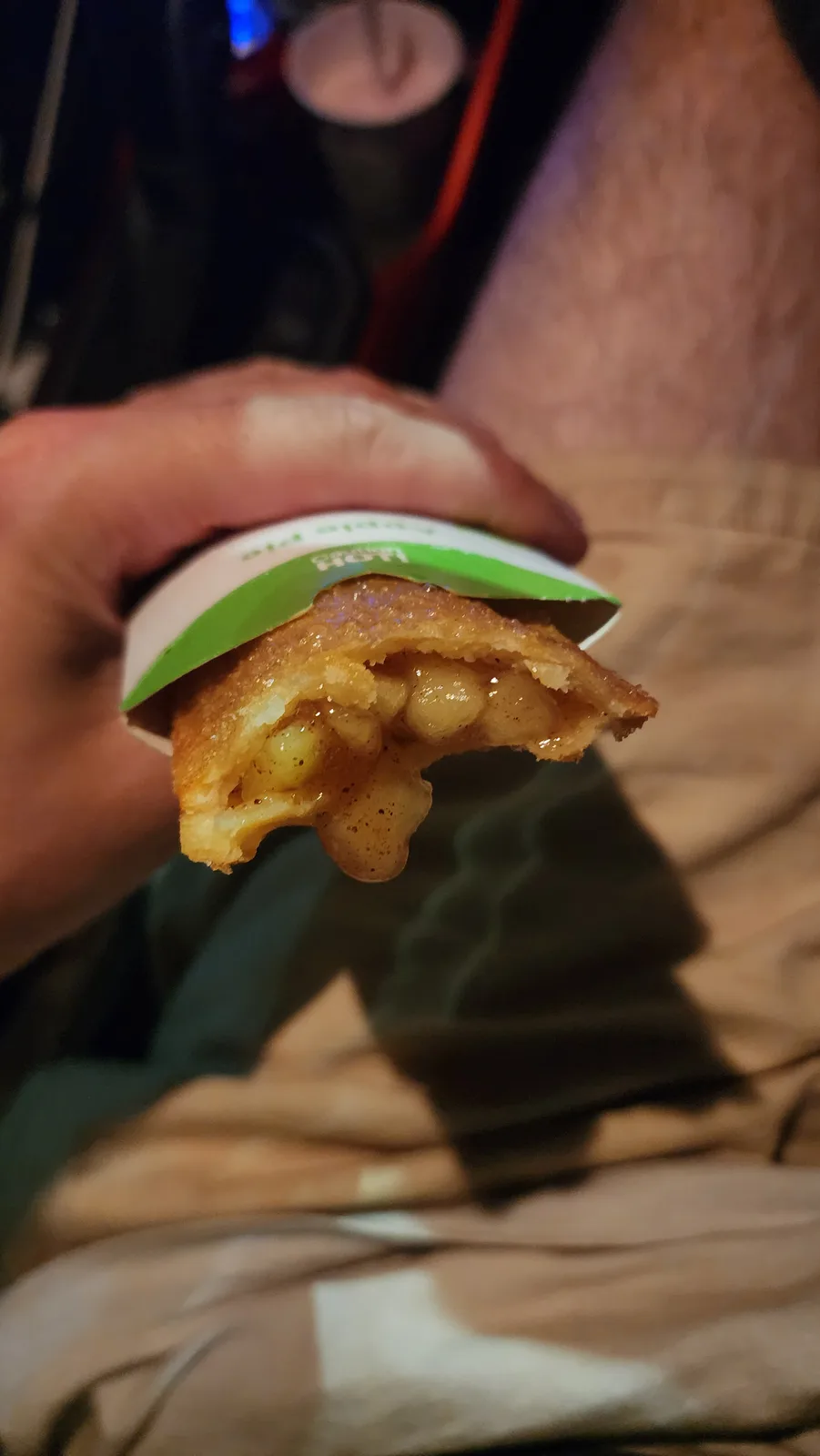 A hand holds a partially eaten, bite-sized apple pie in a white and green wrapper, revealing gooey, chunky apple filling…