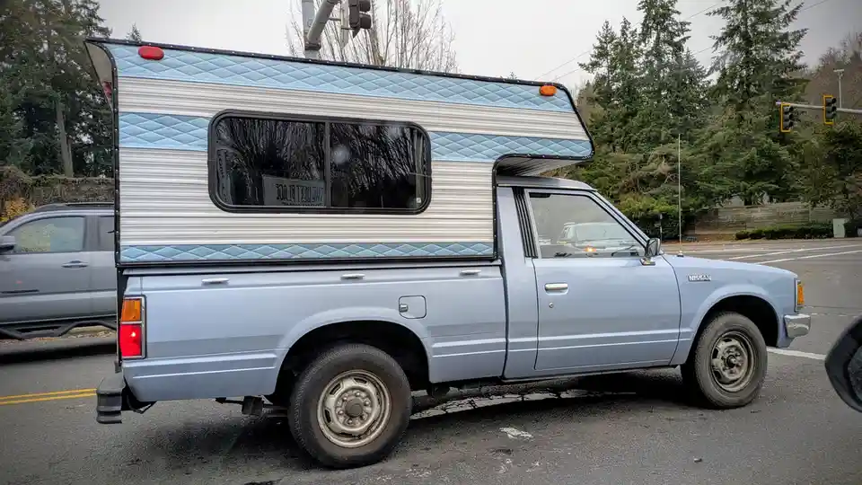 A light blue Nissan pickup truck with a camper shell is stopped at an intersection. The camper shell has a white corrugated…