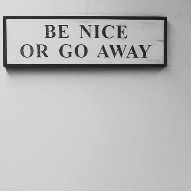 A black-framed white sign with bold black text reading 'BE NICE OR GO AWAY' is mounted on a plain, light-colored wall. The…