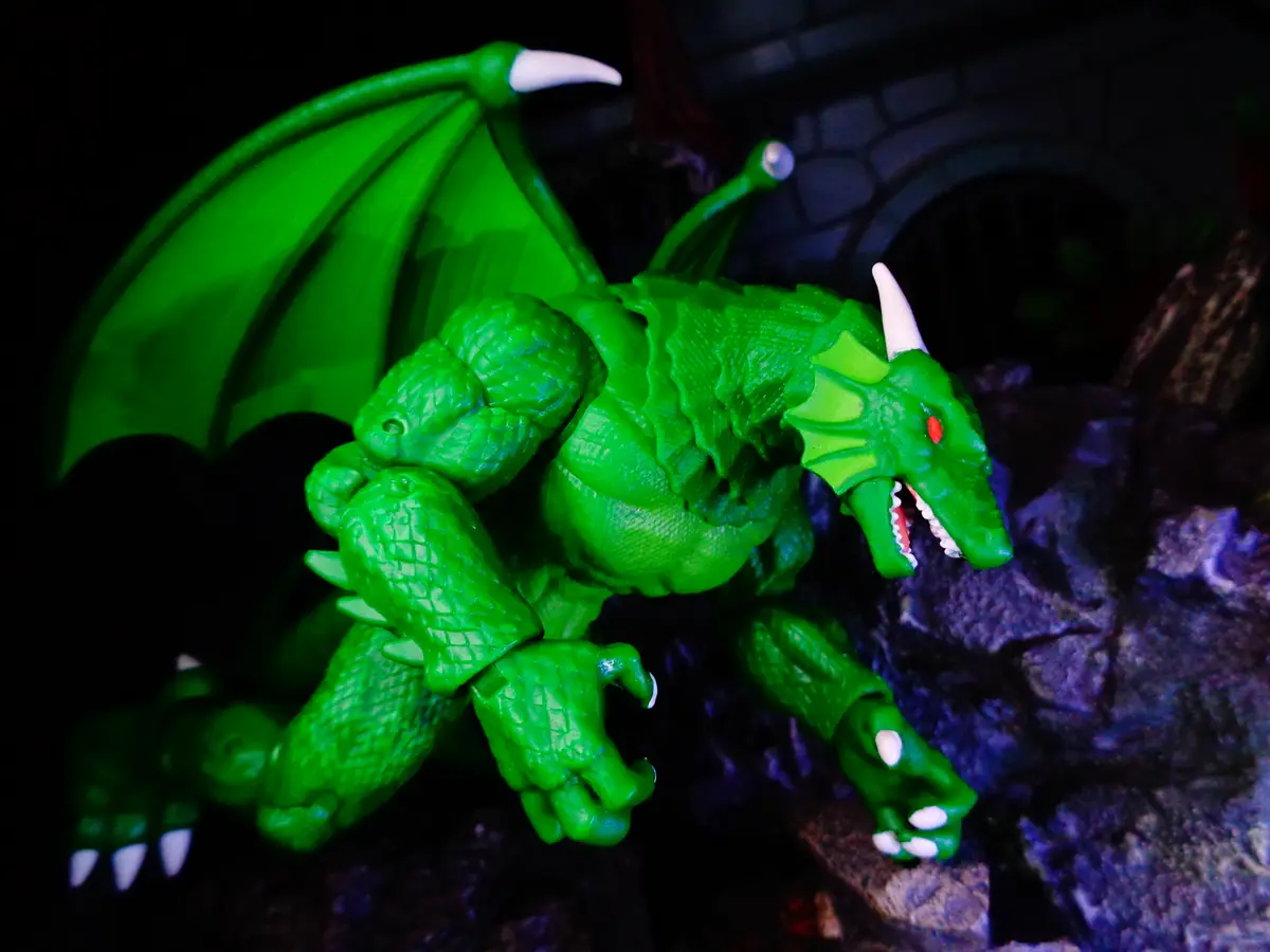 A bright green plastic dragon toy with white claws and horns, red eyes, and spread wings is posed on dark rocks against a…