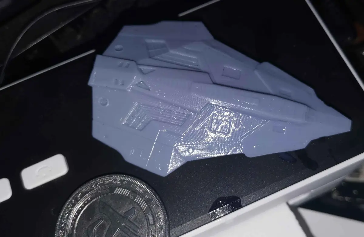 A small, light blue 3d-printed model of a Halo spaceship rests on black foam inside a white box, next to a silver coin and…