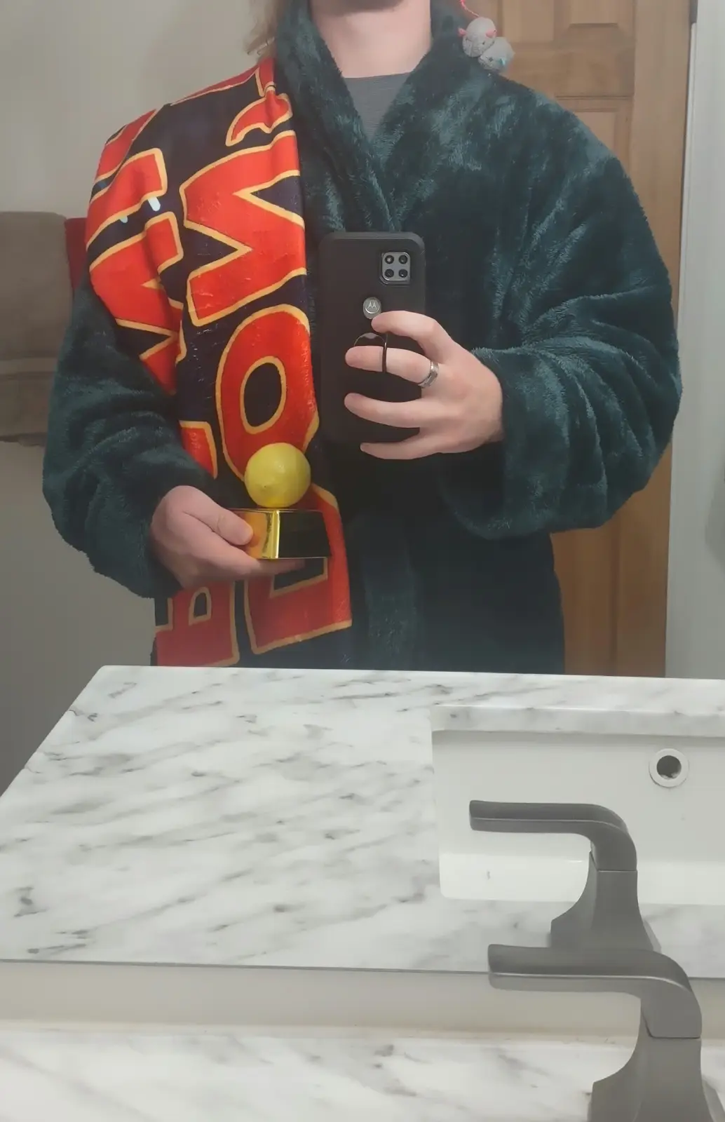 A person in a dark green fuzzy robe and a red and black 'WOW' towel takes a mirror selfie holding a small golden trophy with…