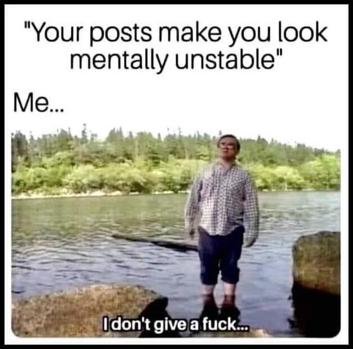A person stands barefoot in shallow water near rocks, wearing a plaid shirt and dark pants, with a forested shoreline behind…