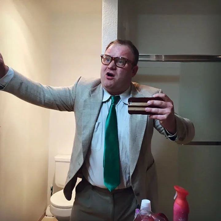 A man in a beige suit, white shirt, and green tie stands in a bathroom, holding a phone to take a selfie in the mirror while…