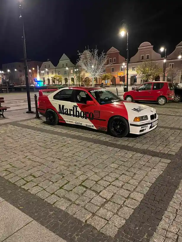 A red and white BMW E36 sedan with prominent Marlboro branding and racing decals is parked on a cobblestone plaza at night…