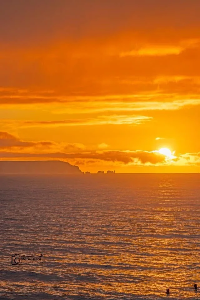 A serene sunset over the ocean with vibrant orange and yellow hues reflecting on the water, a distant landmass on the…