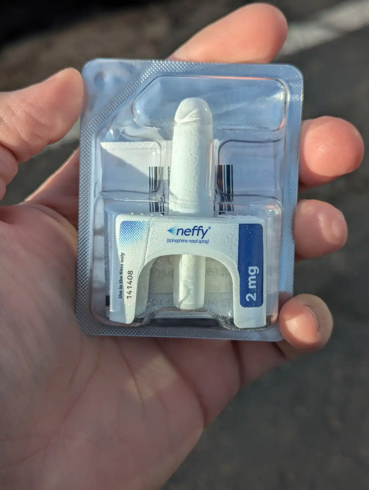 A hand holds a blister pack containing a white, cylindrical neffy epinephrine nasal spray device labeled '2 mg'. The…