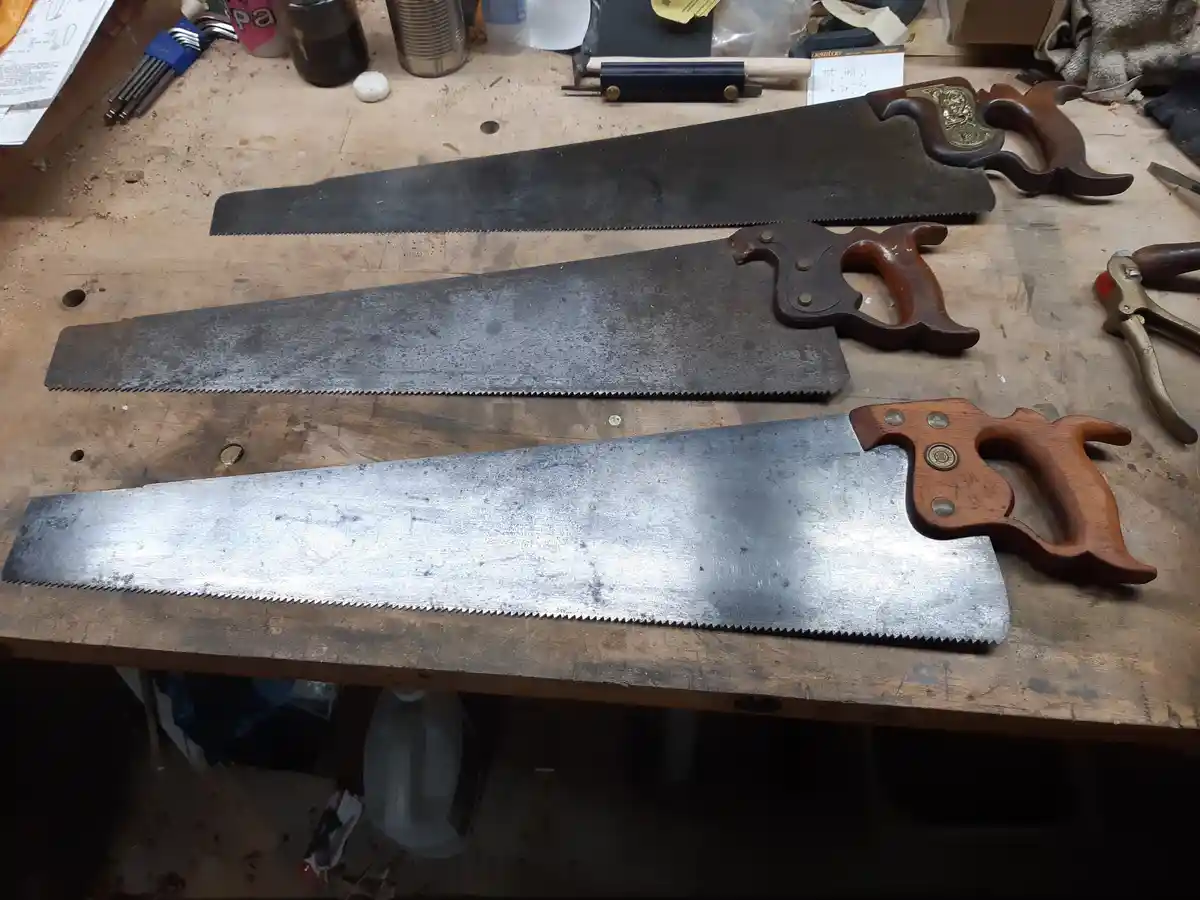 Three hand saws with wooden handles lie on a worn wooden workbench, arranged in descending size from top to bottom. The saws…