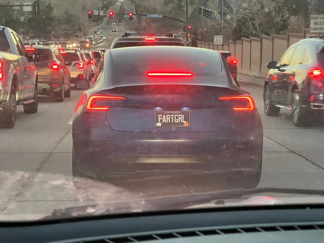 A photo taken from inside a car shows the rear of a dark Tesla Model 3 stopped in traffic with its brake lights on. The…