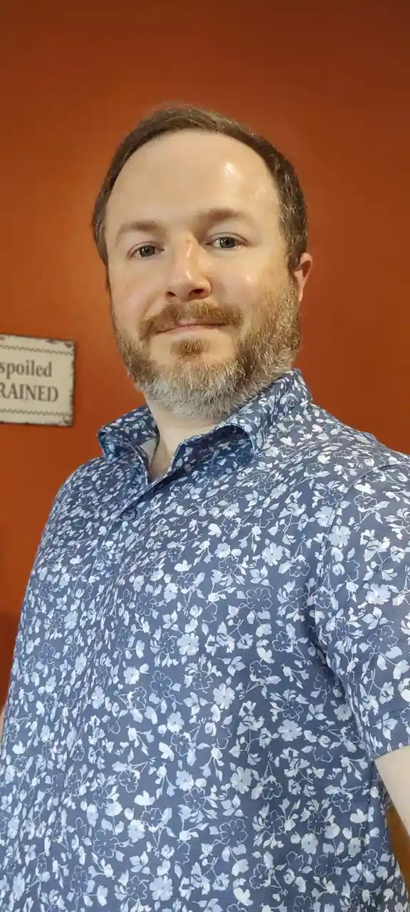 A man with a beard and mustache wearing a blue floral shirt stands in front of an orange wall. A sign behind him reads…