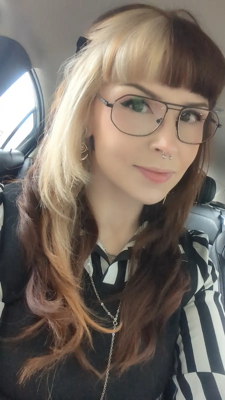 A selfie of a person with two-toned hair (blonde on top, brown underneath) and bangs, wearing glasses and a nose ring…