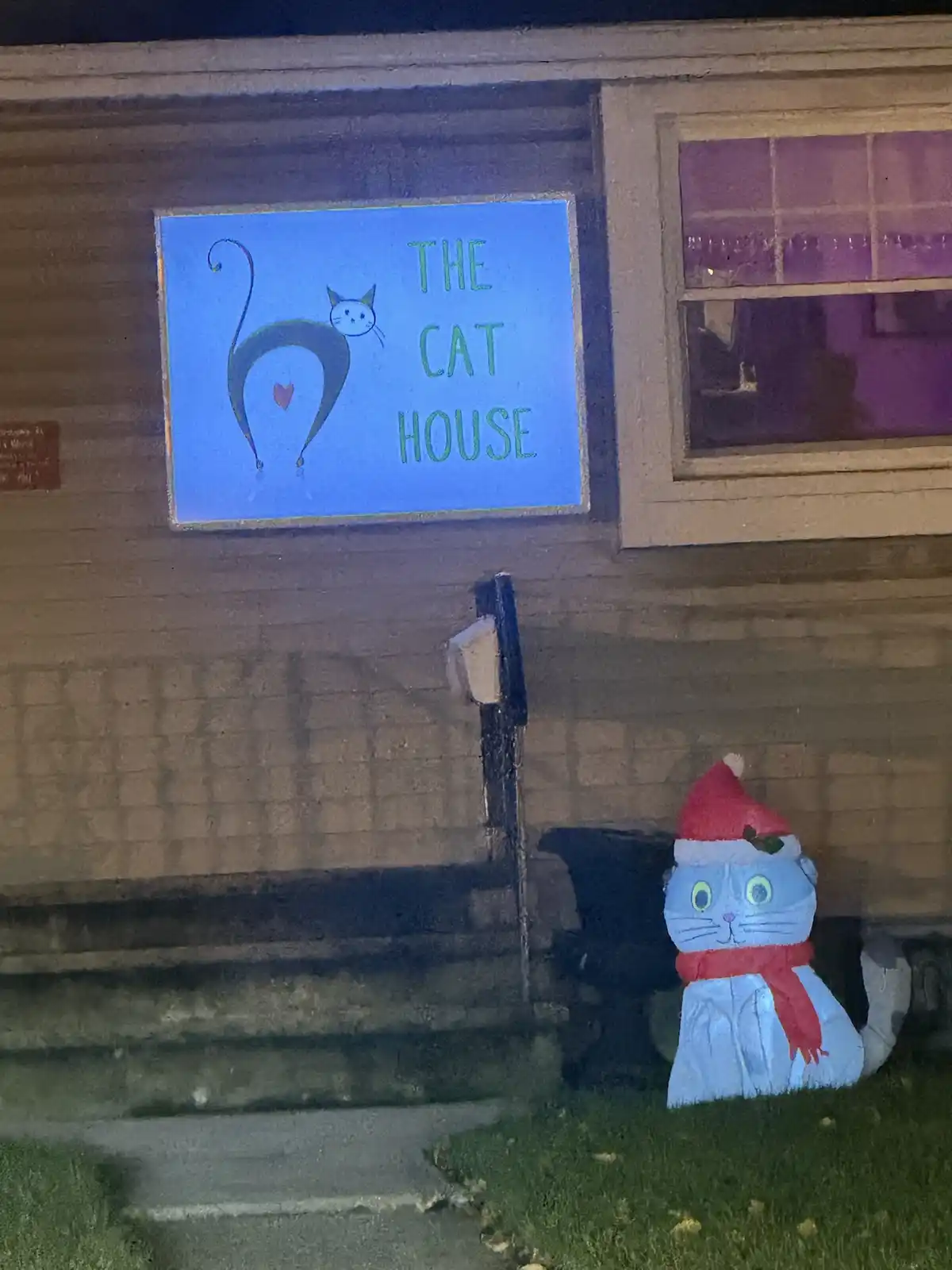 A nighttime photo of a house with a blue sign reading 'THE CAT HOUSE' featuring a stylized cat silhouette with a heart…