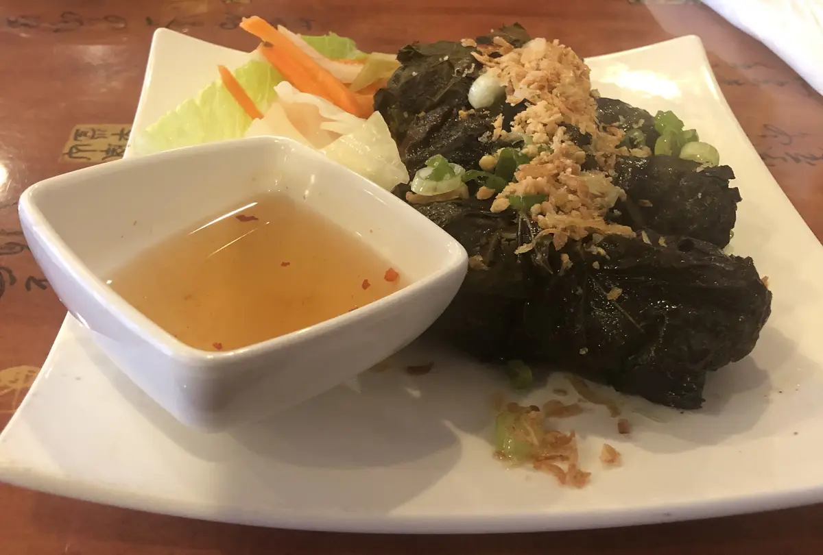 A plate of dark leaf-wrapped food, resembling stuffed leaves or dumplings, topped with fried onions and green onions, served…