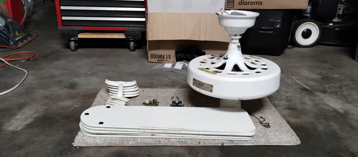 A disassembled white ceiling fan lies on a rug in a garage, with its blades, motor housing, and hardware scattered around…