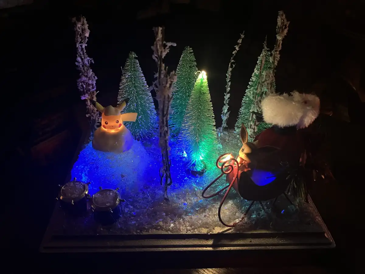 A festive, dimly lit diorama features a glowing Pikachu figurine dressed as a snowman on the left and a Eevee figurine in a…